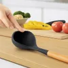 A hand is shown touching a silicone ladle with a wooden handle, placed on a wooden cutting board. In the background, there are vegetables and sliced bell peppers. Náhľad obrázku produktu