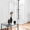A table with decorative items in front of a window with sheer curtains. Produktbild-Vorschau