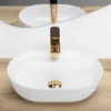A white oval countertop sink with a gold faucet and drain, set against a wooden surface and a mirrored background. Náhled obrázku produktu