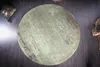 A round rug with a distressed green and brown pattern is laid on a dark wooden floor. A decorative pillow with a gold leaf pattern is visible in the top left corner, and a textured decorative object is in the top right corner. Produktbild-Vorschau