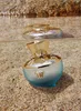 A perfume bottle sits on a sandy beach with its reflection visible in the water. Anteprima dell’immagine prodotto