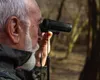 An elderly man looking through binoculars in a forest. Podgląd zdjęcia produktu