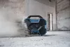 A blue and black Bosch radio is on a concrete floor in a construction site, with dust blowing around it. Produktbild-Vorschau