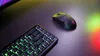 A black wireless gaming mouse and a black mechanical keyboard with green backlighting are placed on a dark surface. The background is illuminated with colorful lights. Produktbild-Vorschau