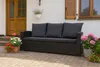 A black outdoor sofa with dark gray cushions is placed on a paved patio in front of a white house with wooden doors and shutters. Potted plants with pink, red, and white flowers are arranged around the sofa. Anteprima dell’immagine prodotto