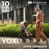 A woman pushes a baby stroller with a child in it, walking on a paved path next to a building and greenery. Produktbild-Vorschau