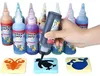 Eine Hand drückt eine schwarze Aqua Fairy Farbflasche auf eine Pinguin-Schablone, mit anderen bunten Farbflaschen und Schablonen im Hintergrund Produktbild-Vorschau
