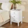 A white nightstand with three drawers is placed next to a bed. A vase with greenery sits on top of the nightstand. Podgląd zdjęcia produktu