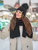 A woman wearing a winter hat and scarf smiles in a snowy outdoor setting with festive lights and buildings in the background. Produktbild-Vorschau