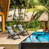 Two grey lounge chairs are set up on a wooden deck next to a swimming pool, surrounded by lush tropical vegetation and palm trees. Voir les images du produit