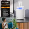 A lifestyle image shows a woman relaxing on a couch with a mug, while a white aroma diffuser with blue LED lights emits steam in the background, placed on a wooden-accented table. A black panel on the left lists the benefits of the aroma diffuser. Produktbild-Vorschau