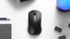 A black wireless mouse is centered on a white surface, with a pencil and a notebook to the left, and Lego bricks and a glowing blue object to the right. Voir les images du produit