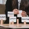 A person is holding a blank card in front of several cards with words like 'MANAGEMENT', 'SUPPORT', and 'MARKETING' written on them, which are placed on wooden blocks. Voir les images du produit
