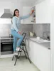 A woman is using a dark gray, 3-step folding ladder to reach items in a kitchen cabinet. Produktbild-Vorschau