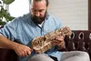 A man is sitting on a brown leather couch, holding and examining a wooden 3D puzzle. Produktbild-Vorschau