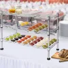 A chrome shelving unit with multiple tiers filled with small bowls of various dips and sauces, set up on a table with a white tablecloth. In the background, a blurred catering setup is visible. Produktbild-Vorschau