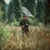 A man wearing a jacket and hat walks through a forest with a pair of binoculars in his hands. There is a small wooden cabin in the background. Produktbild-Vorschau