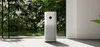 A white air purifier is placed on a light gray floor in front of a large window with lush green plants visible outside. Produktbild-Vorschau