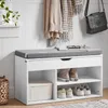 A white shoe bench with a gray cushion, featuring storage compartments for shoes and a drawer. It is placed against a white paneled wall, with clothes and a handbag hanging on hooks above. Anteprima dell’immagine prodotto