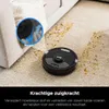 A robot vacuum cleaner is cleaning a carpet with dirt on it, next to a sofa. A close-up inset shows the vacuum's cleaning mechanism. Produktbild-Vorschau