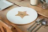 A round placemat with a white surface and a wooden star design in the center, set on a wooden table with cutlery, a candle, and a vase. Podgląd zdjęcia produktu