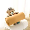 A teddy bear wearing a hat holds a rolled-up blanket on a white table. Produktbild-Vorschau
