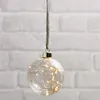 A clear glass ornament filled with fairy lights hangs against a white brick wall. Produktbild-Vorschau