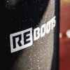 A close-up of a black product with a white logo, showing water droplets on its surface. The background is blurred and appears to be a reddish-brown surface. Produktbild-Vorschau