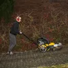 A person is using a wildkrautbürste (weed brush) to clean a paved area next to a garden with bushes. Produktbild-Vorschau