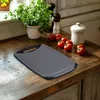 A cutting board is placed on a wooden countertop next to tomatoes and a potted plant. Anteprima dell’immagine prodotto
