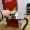 A person's hand is guiding a piece of wood through a red bandsaw. The bandsaw is on a wooden surface, and there is a black hose attached to it. Produktbild-Vorschau