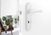 A white door handle and lock mechanism are shown on a white door, with a blurred interior scene in the background. Produktbild-Vorschau