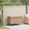 A beige poly rattan garden chest with wheels is placed on a patio next to a wooden fence. Podgląd zdjęcia produktu