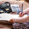 A child is drawing in a sketchbook with a pencil, surrounded by art supplies. Podgląd zdjęcia produktu