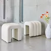 Two white upholstered stools are placed in a modern interior with concrete walls and a concrete floor. A vase with orange flowers is on the right. Produktbild-Vorschau