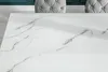A close-up view of a white marble-patterned tabletop with two tufted gray chairs visible in the background. Náhled obrázku produktu