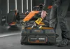 A person is placing a yellow power tool into a black and orange tool bag, with a race car in the background. Produktbild-Vorschau