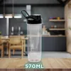 A clear shaker bottle with a black lid and handle sits on a wooden surface in front of a blurred kitchen background. Produktbild-Vorschau
