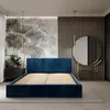 A bedroom scene featuring a dark blue upholstered bed frame with a slatted base, set against a wall with a nature-themed mural and vertical wooden slats. A round mirror and a bedside table with flowers are also visible. Produktbild-Vorschau