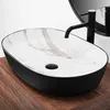 A modern oval countertop sink with a black exterior and a white marble-patterned interior, featuring a black faucet and drain, placed on a wooden surface. Náhľad obrázku produktu
