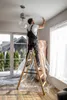 A man is painting a ceiling while standing on a wooden ladder. Produktbild-Vorschau