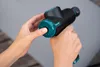 A person's hands are holding a black and teal massage gun. One hand is gripping the handle, while the other is pressing a button on the head of the device. Produktbild-Vorschau
