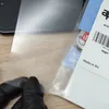 A person wearing black gloves is holding a clear protective film over a wooden surface. Packaging with text and a barcode is visible to the right. Produktbild-Vorschau