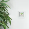 A small white framed picture with a tree and text is on a white wall, with a plant in the foreground. Produktbild-Vorschau
