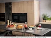 A modern kitchen scene with a table set for a meal, featuring built-in ovens in the background. Produktbild-Vorschau