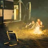 A couple is sitting by a campfire next to a van at night, with a portable LED spotlight illuminating the scene. Produktbild-Vorschau