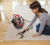 A woman is kneeling on a carpet and using a portable carpet cleaner to clean a stain. Voir les images du produit