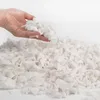 A hand holding a pile of white foam flakes over a larger pile of the same material. Produktbild-Vorschau