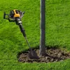 A gas-powered auger is positioned next to a tree trunk in a grassy yard. Podgląd zdjęcia produktu