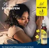 A woman sleeping peacefully in bed, with a product bottle and flowers placed on her arm. Produktbild-Vorschau